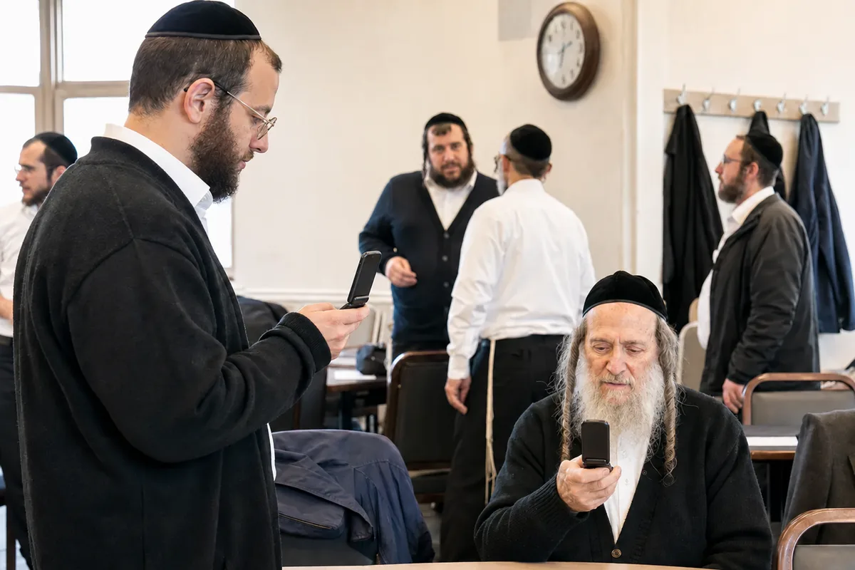 Men in a shul between minyanim, one standing and texting on a flip phone while a seated man reads a message on his.